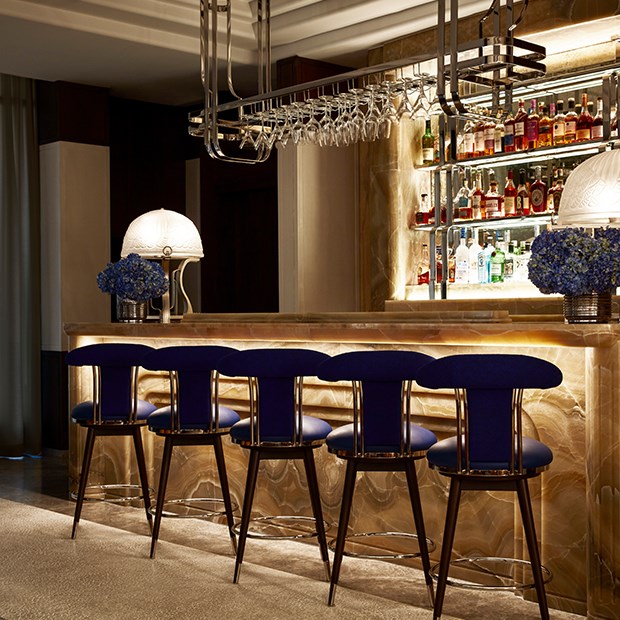 Five dark blue bar stools sitting at The Maybourne Bar. Bottles of a various alcohol brands are on display behind the bar