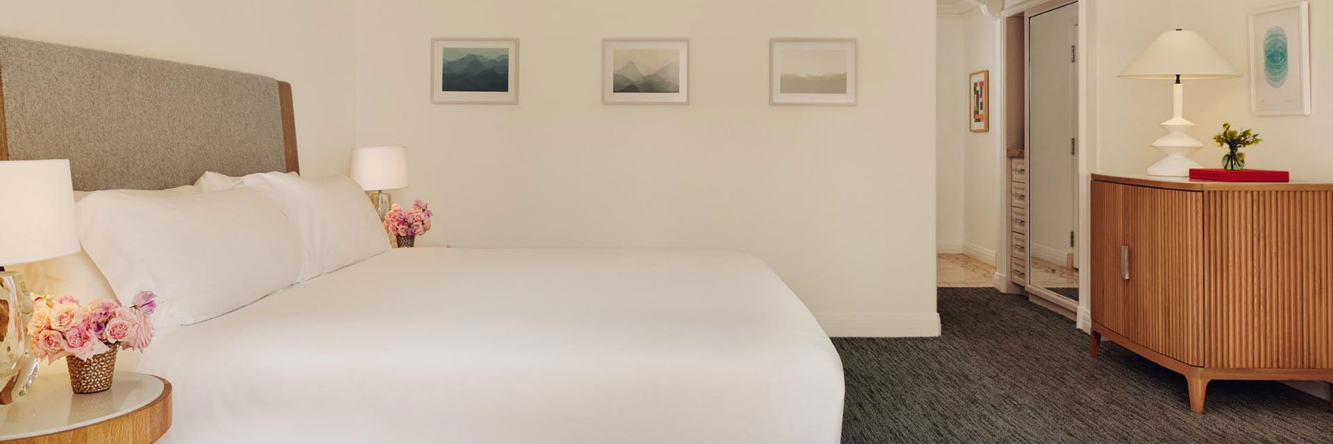 A well-lit bedroom with a minimalist design. A white double bed with a neutral headboard sits to the left. There are nightstands with matching table lamps and vases filled with pink flowers on either side. There is a wooden dresser to the right and a table lamp and small plant on top. The floor is covered with a carpet.