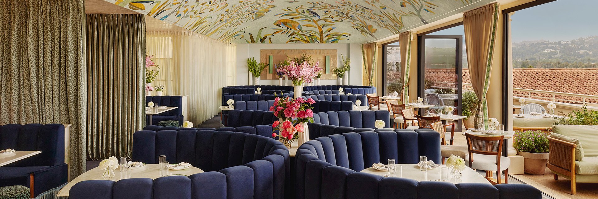 Dark blue booths surrounding tables with small pink roses on them, on the rooftop bar Dante with a ceiling mural above.