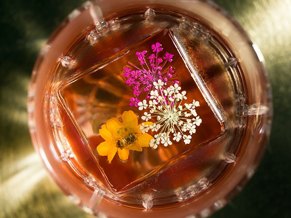 A top down view of a cocktail glass with a large cube of ice. Inside the ice are yellow, white, and pink flower petals.