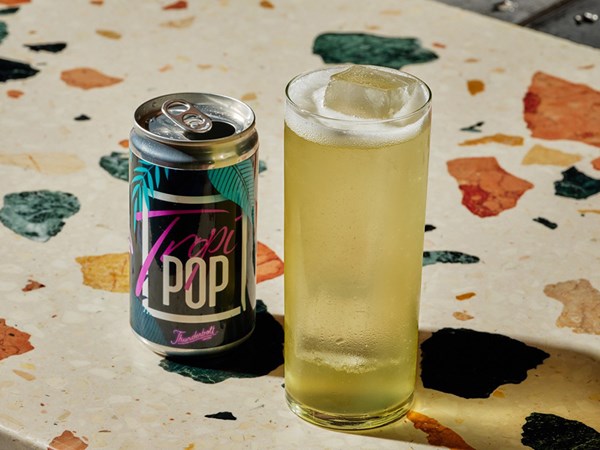 A can of Thunderbolt Tropi Pop. To the right of the can is a tall cocktail glass filled with a yellow drink.
