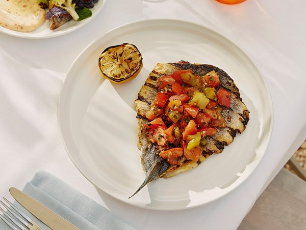 A grilled branzino with heirloom tomatoes plated on a table with a white tablecloth. A lemon cut in half and charred.