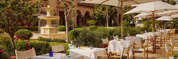 Outside dining tables and chairs with white umbrellas. Behind, there's a fountain in a garden surrounded by bushes and tree.