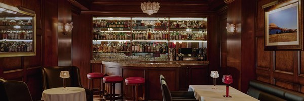 A dimly lit marble bar with red stools. Whisky brands line the glass shelves behind the bar. There are tables with lamps.