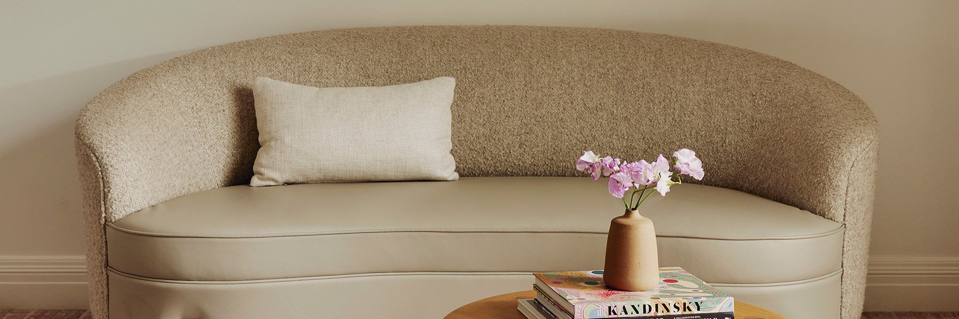 A curved ivory couch with a small, white pillow. A stem with pink flowers is in a vase on top of a stack of book on a table.