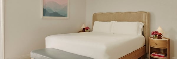 A king sized bed with white sheets. Two nightstands with lamps and violet potted roses.