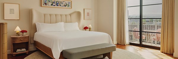 A king sized bed with white sheets. Two nightstands with red flowers. Glass doors lead to a terrace with pink furniture.