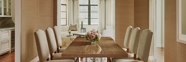 A dining room table with six chairs and pink flowers. There's a walkway to a living area with a couch, table, and lamp.