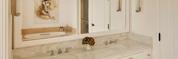 A marble countertop with two sinks. In the mirror you can see a bathtub and a painting of person.
