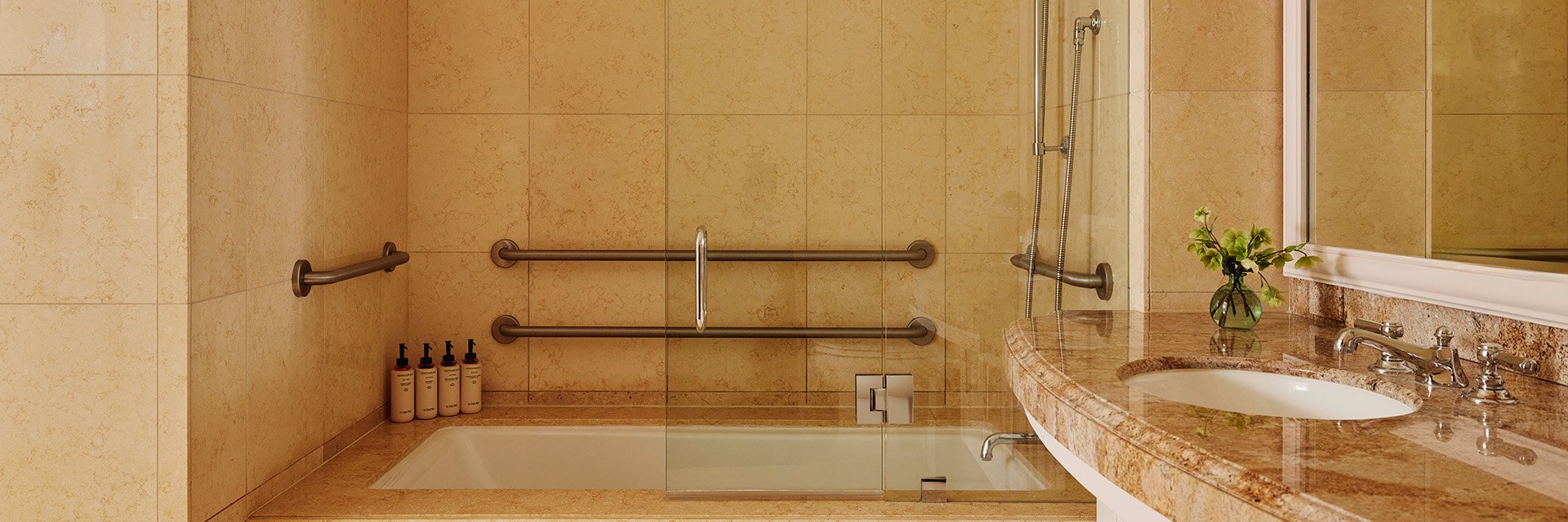 An accessible bathtub with handrail in a brown marble bathroom with mosaic wall tiles.