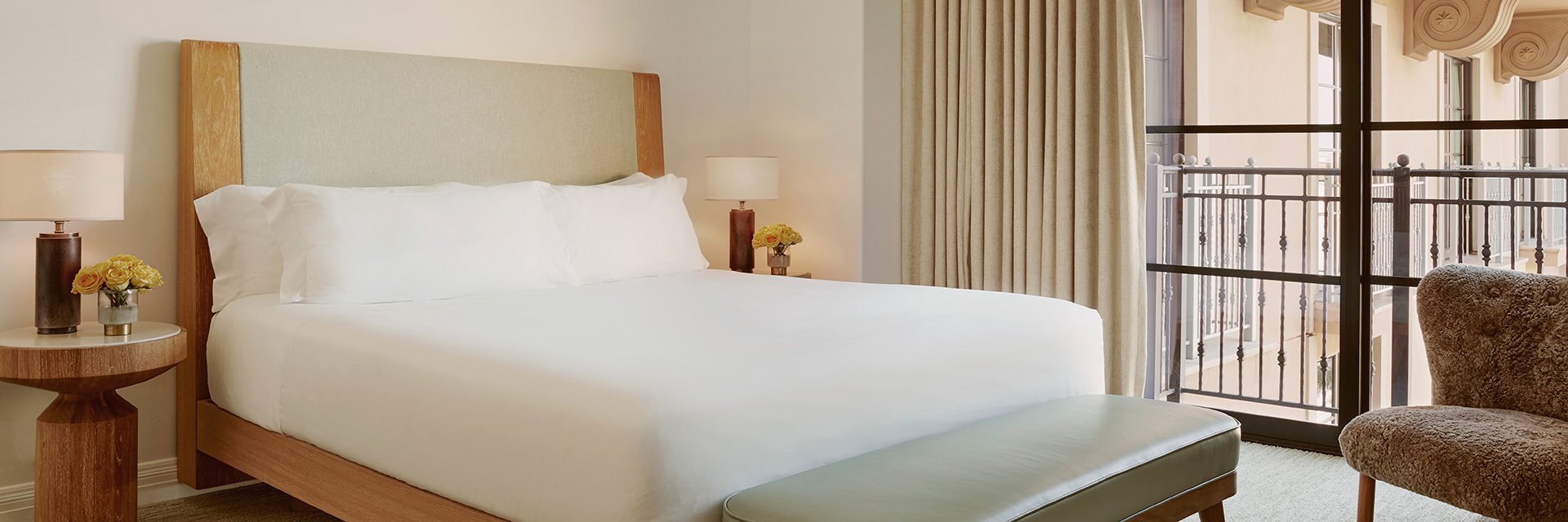 A king sized bed with white sheets in a hotel bedroom. There are yellow roses in glass vases on nightstands and a window.