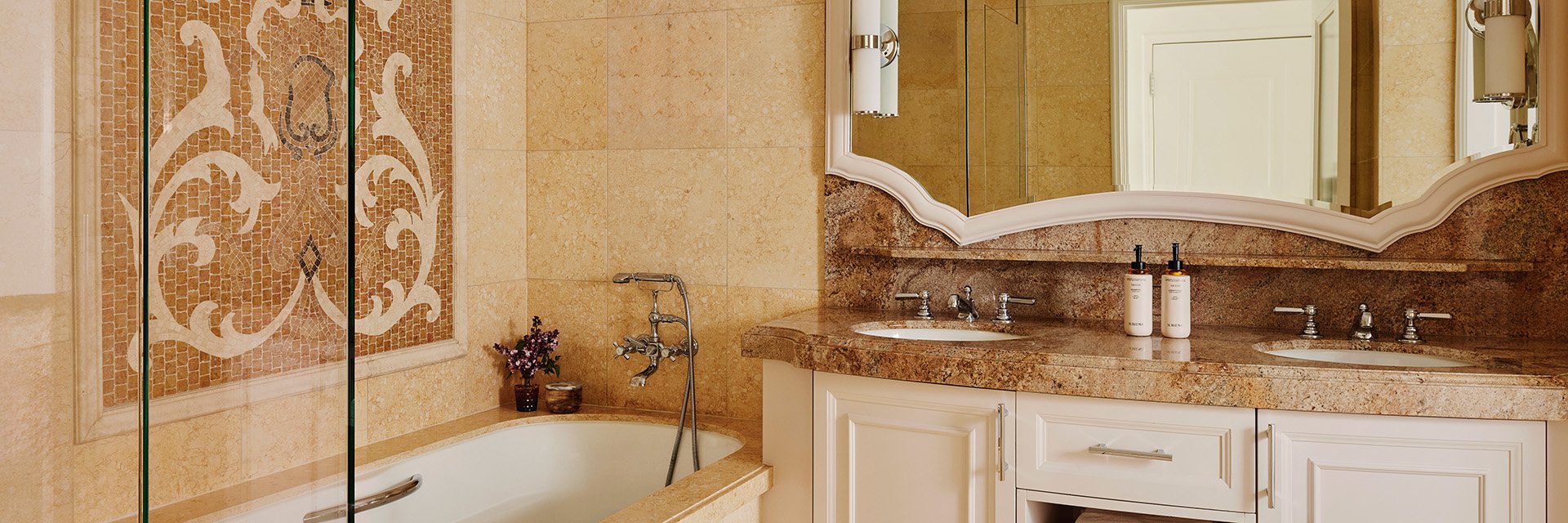 A tiled bathroom, a bathtub with a tiled wall mosaic, and a brown marble sink with white drawers.
