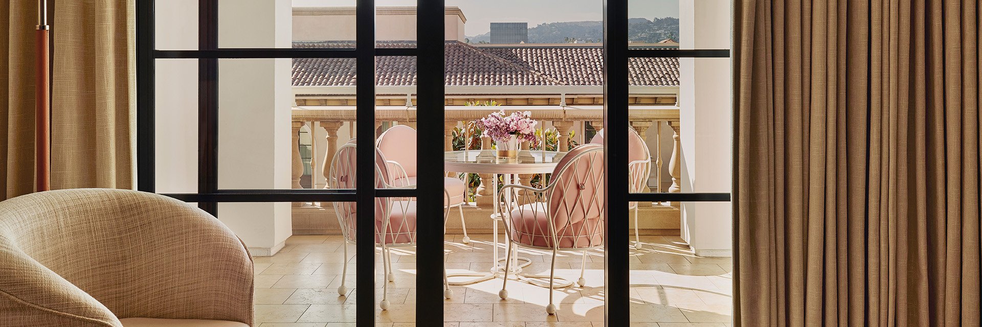 Open sliding glass doors that show an ivory tiled terrace with a glass table and pink potted flowers with four pink chairs.