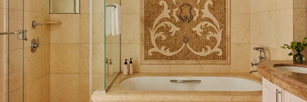 A tiled bathroom, a bathtub with a tiled wall mosaic, a hanging white towel, and a brown marble sink with white drawers.