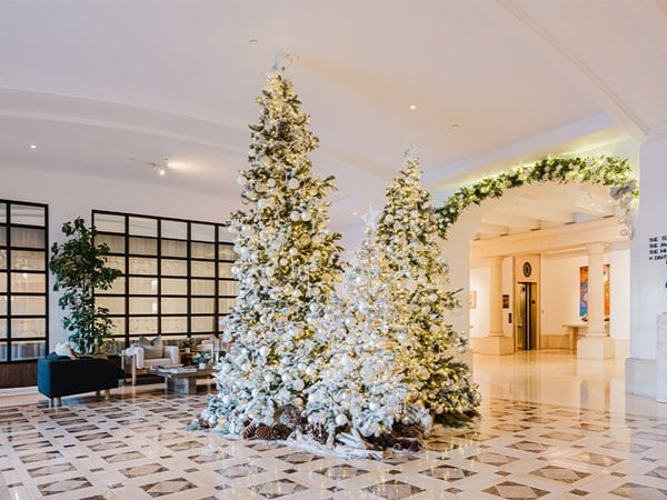 A festive display of snow-dusted Christmas trees decorated with shimmering white ornaments and warm lights, situated in the spacious and elegant lobby of a luxury hotel.