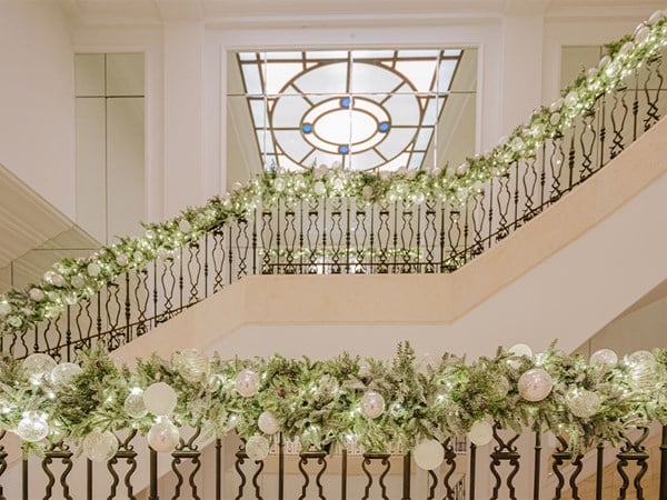 A grand staircase adorned with lush holiday garlands featuring sparkling white ornaments and delicate lights, under a stunning stained glass ceiling.