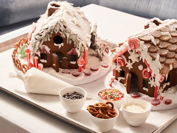 Decorated gingerbread houses with snow-like frosting, red and white candy decorations, and bowls of sprinkles, pretzels, and chocolate chips ready for assembly.