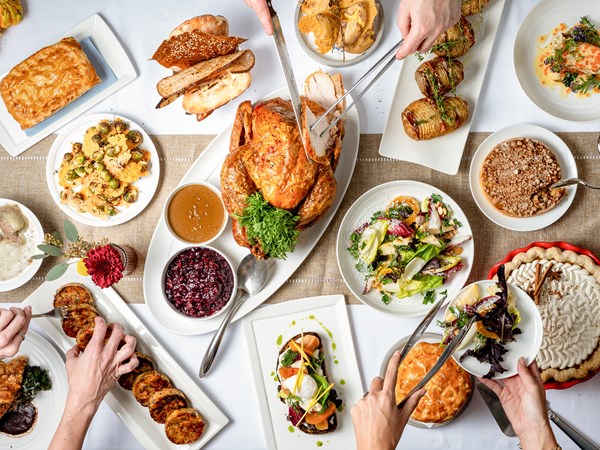 A festive Thanksgiving spread featuring a roasted turkey, cranberry sauce, various sides, breads, and desserts, with hands serving food from the dishes on a decorated table.