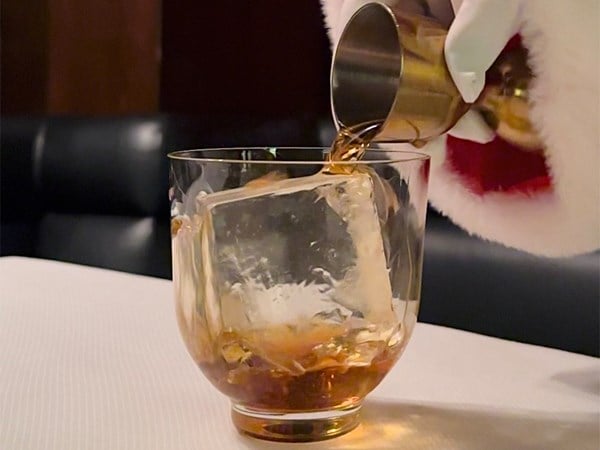 A close-up of whisky being poured over a large crystal-clear ice cube into a glass, evoking luxury and refinement. A gloved hand holding a festive red velvet pouch adorned with a sleek ribbon, against a backdrop of holiday decorations.