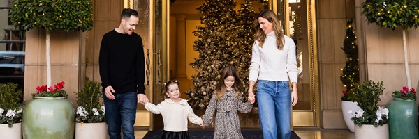 A family of four walks hand-in-hand outside a festive building with a grand Christmas tree glowing in the background.