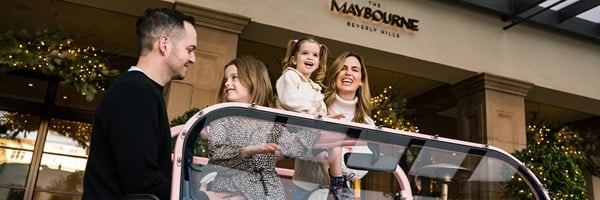 A smiling family enjoys holiday cheer in front of The Maybourne Beverly Hills, decorated with festive greenery and lights.