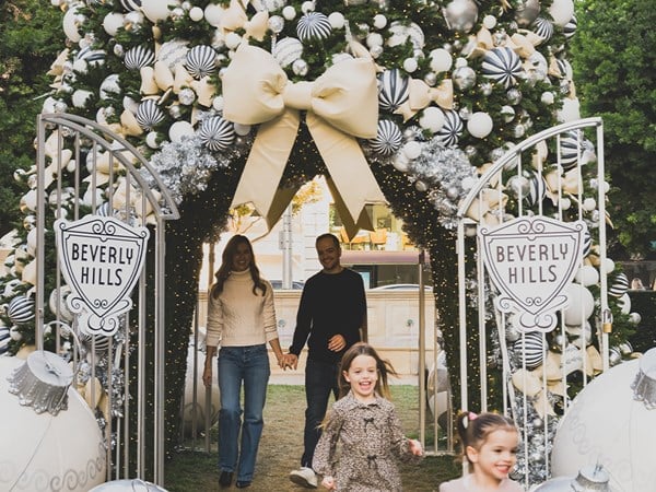 A family walks through a festive, elegantly decorated holiday archway in Beverly Hills, adorned with oversized ornaments, ribbons, and glowing lights.